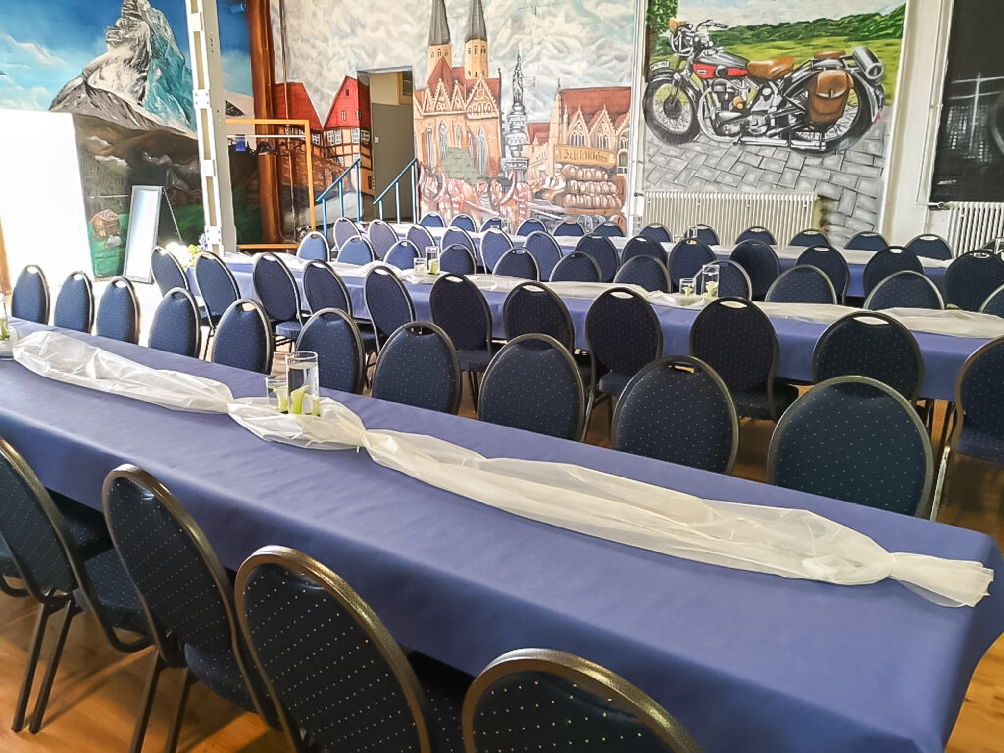 Stimmungsvoll gedeckter Saal mit blauer Tischdekoration und künstlerischer Wandgestaltung – ideal für Feiern, Hochzeiten und Jubiläen.