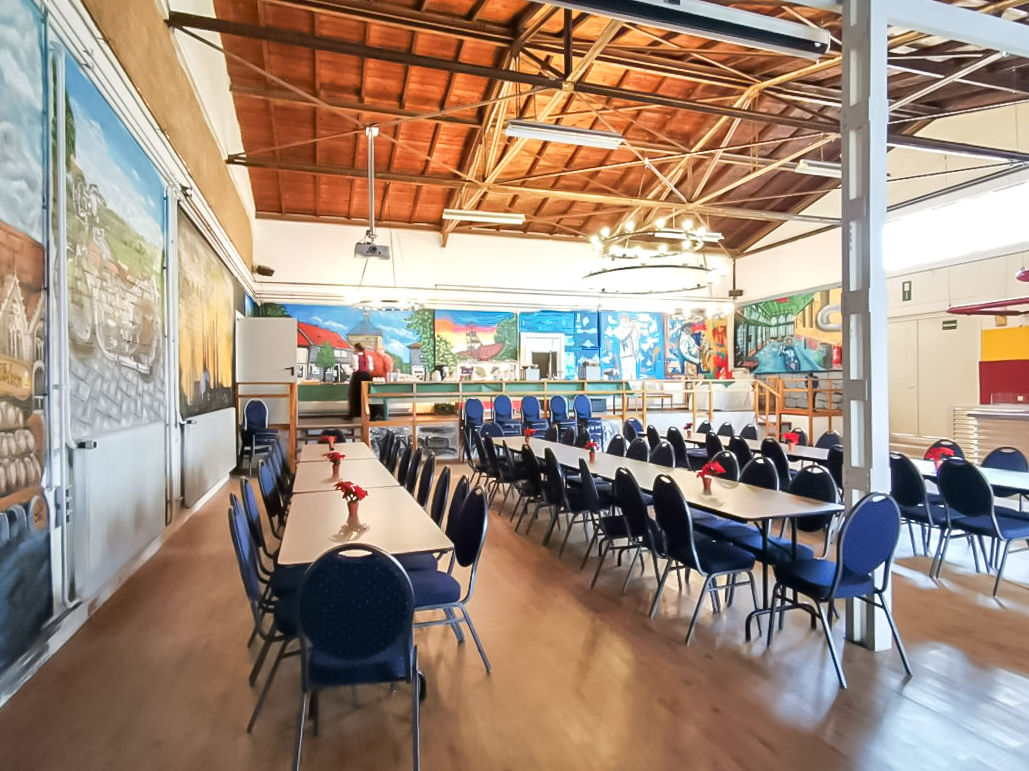 Blick in die weitläufige Meisterhalle mit festlich gedeckten Tischen, bunten Wandbildern und einem integrierten Thekenbereich.
