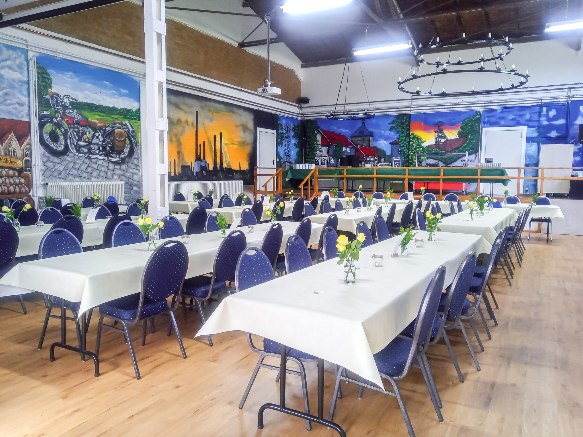 Festlich gedeckter Saal mit gelb-grüner Tischdekoration, farbenfrohen Wandmalereien und Bühne für Feiern und Events.