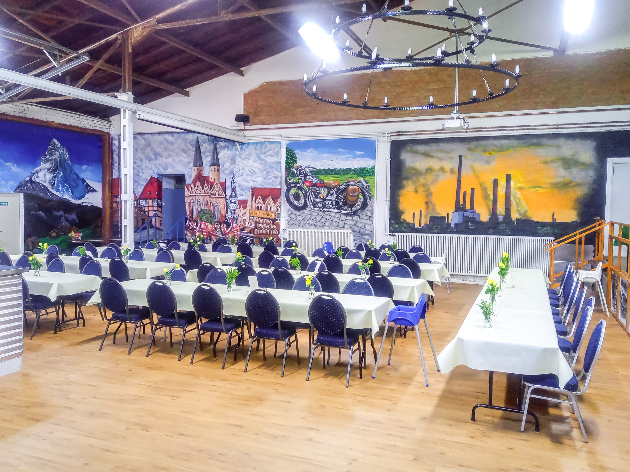 Weitläufige Meisterhalle mit festlich gedeckten Tischen, künstlerischer Wandgestaltung und Bühne für Feste, Konzerte und Firmenfeiern.