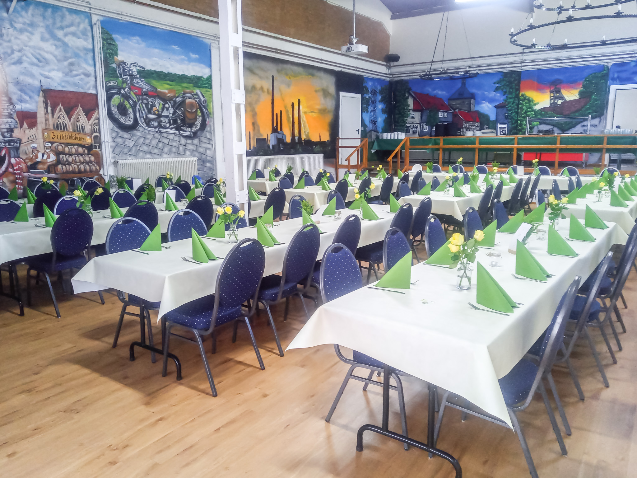 Eingedeckte Meisterhalle mit grüner Tischdekoration, Bühne und farbenfroher Wandmalerei für Feste und Veranstaltungen.